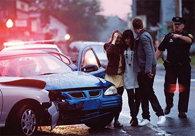 in the early morning, injured young people stand at the site of a car crash as a cop looks on