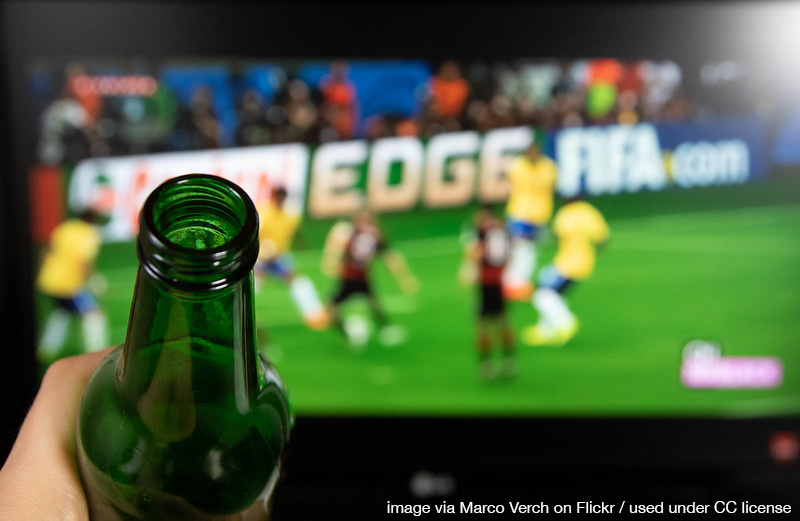 a hand holds a beer bottle up in front of an ongoing soccer game